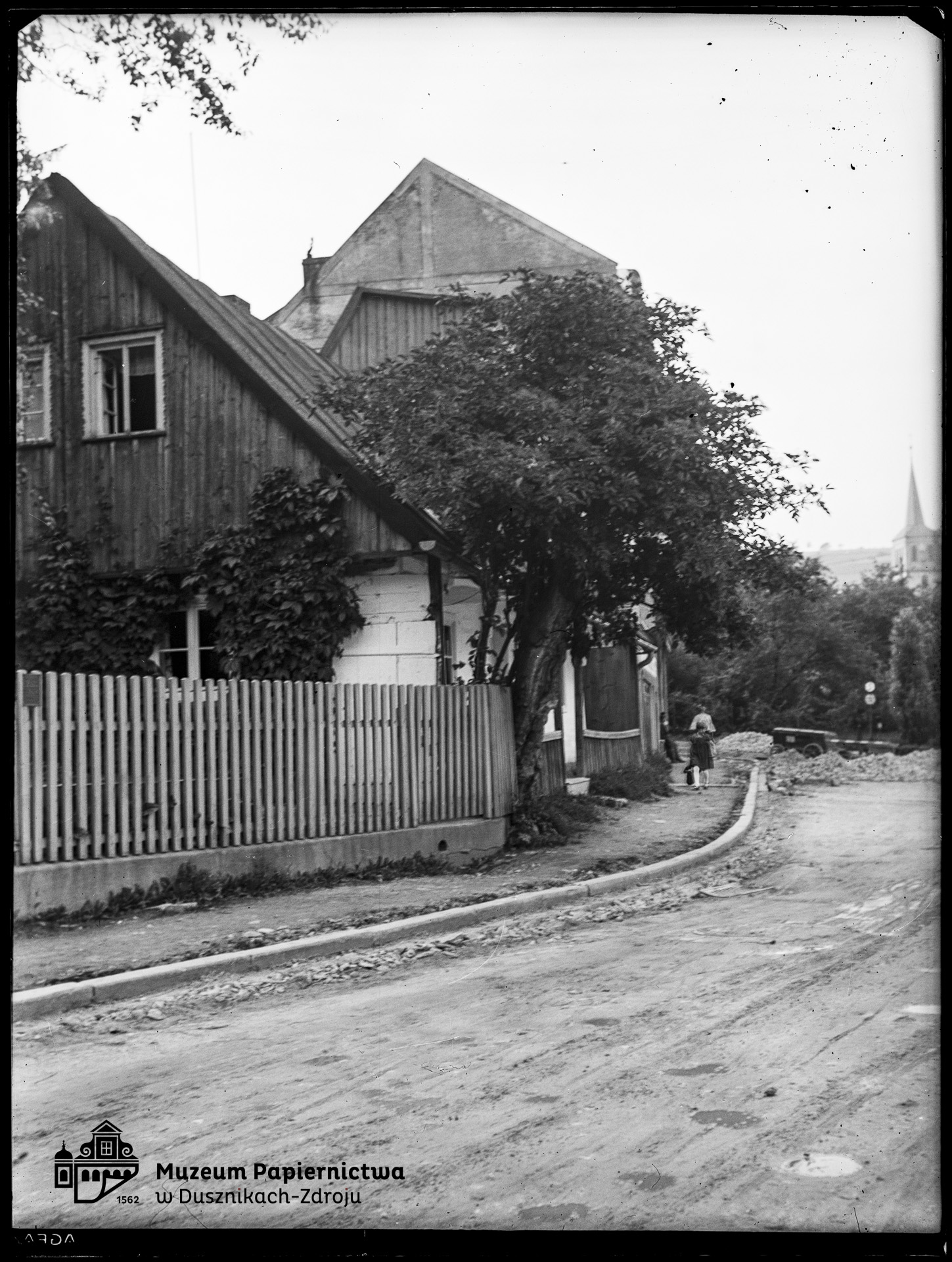 Zdjęcie przedstawiające dom, drogę i w oddali wieżę kościoła pw. św. św. Piotra i Pawła w Bad Reinerz (Duszniki-Zdrój)
